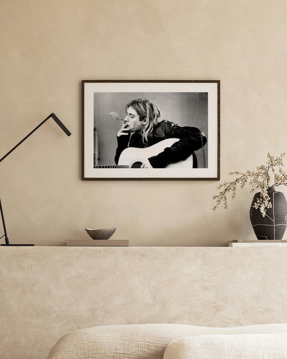 Affiche noir et blanc de Kurt Cobain fumant, guitare en main, au-dessus dune table avec vase et livres.