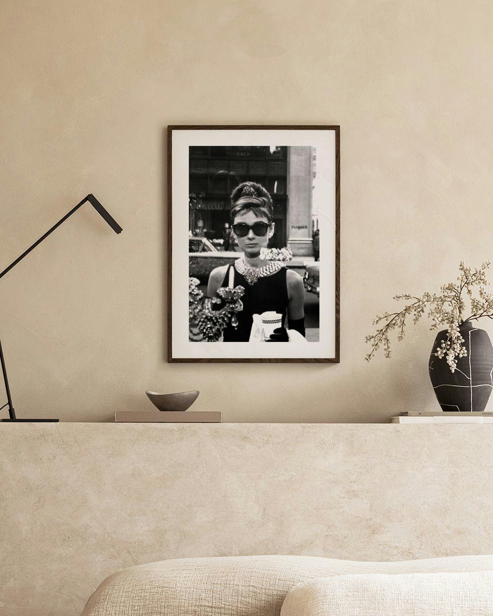 Affiche noir et blanc dAudrey Hepburn avec lunettes de soleil, robe noire et collier, accrochée au-dessus dune étagère moderne.