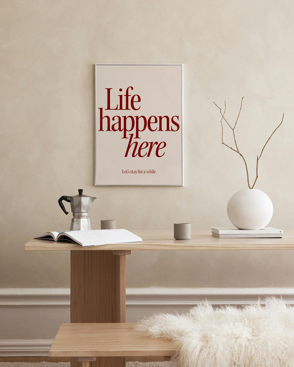 Affiche Life happens here en lettres bordeaux sur fond beige, accrochée au-dessus dune table en bois clair dans un intérieur mod