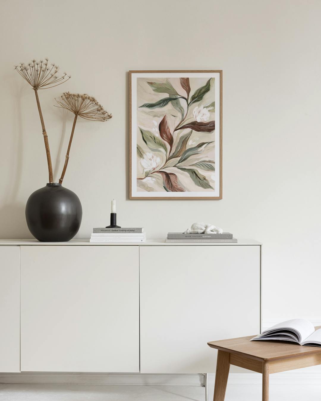 Affiche botanique avec des feuilles vertes et marron et des fleurs blanches, suspendue au-dessus dun buffet blanc avec un vase.